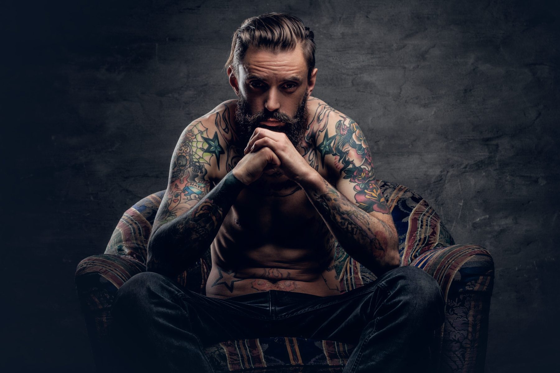 bearded male with naked tattooed torso. Portrait of a thoughtful bearded male with tattooed naked torso and crossed arms, sits on a chair over dark grey background.